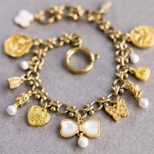 Puede incluir: Pulsera de abalorios dorada con varios dijes, incluyendo un corazón, una mariposa y un lazo. La pulsera tiene un cierre de palanca y está adornada con perlas de imitación y un dije de trébol de cuatro hojas. La pulsera se muestra sobre una superficie gris.