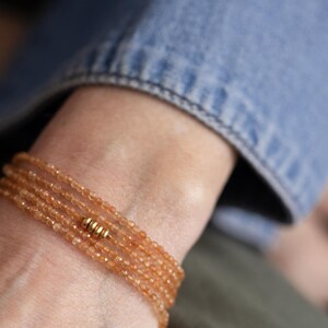 Op de afbeelding: Een armband met meerdere strengen met kleine, gefacetteerde oranje kralen. De armband is geaccentueerd met goudkleurige metalen kralen. De armband wordt om de pols gedragen, met een spijkeroverhemdmouw op de achtergrond.