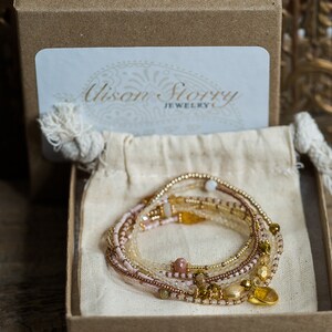 May include: A set of multi-strand beaded bracelets in shades of pink, gold, and brown, with a gold charm in the shape of a heart. The bracelets are displayed in a small, brown box with a white label that reads "Alison Story Jewelry".