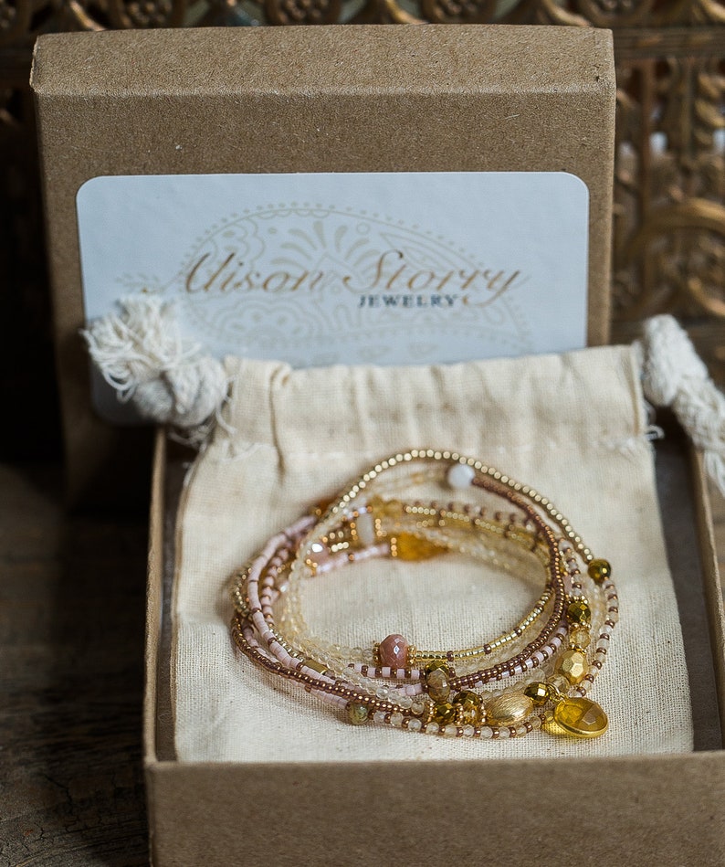 May include: A set of multi-strand beaded bracelets in shades of pink, gold, and brown, with a gold charm in the shape of a heart. The bracelets are displayed in a small, brown box with a white label that reads "Alison Story Jewelry".