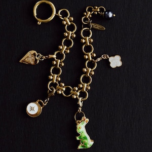 Puede incluir: Pulsera de abalorios dorada con varios dijes, incluyendo un coraz&oacute;n, un tr&eacute;bol de cuatro hojas, un conejo y un colgante tipo br&uacute;jula. La pulsera tiene un cierre de mosquet&oacute;n y una etiqueta peque&ntilde;a que dice "Alison Berry Jewelry".