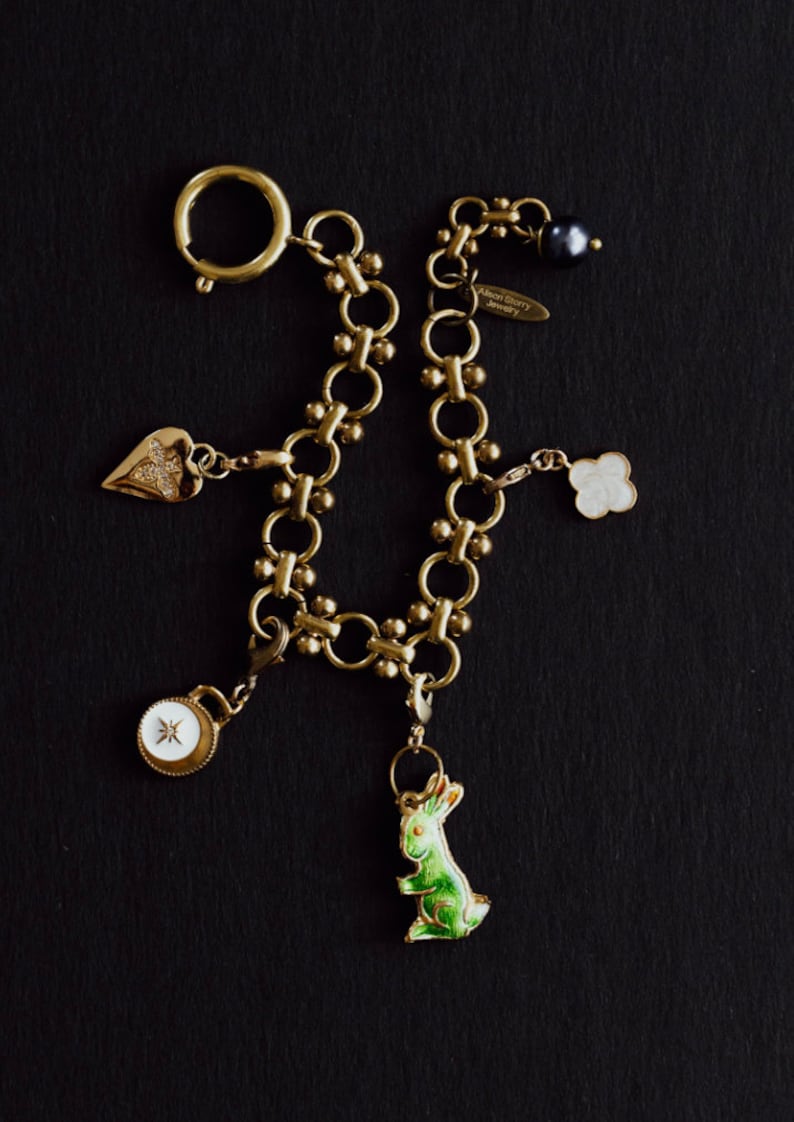 Puede incluir: Pulsera de abalorios dorada con varios dijes, incluyendo un coraz&oacute;n, un tr&eacute;bol de cuatro hojas, un conejo y un colgante tipo br&uacute;jula. La pulsera tiene un cierre de mosquet&oacute;n y una etiqueta peque&ntilde;a que dice "Alison Berry Jewelry".