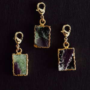May include: Three gold-colored charm pendants with rectangular, raw-edged stone insets. The stones have a mix of green, purple, and white hues, and are edged with gold. Each charm has a gold-colored clasp.