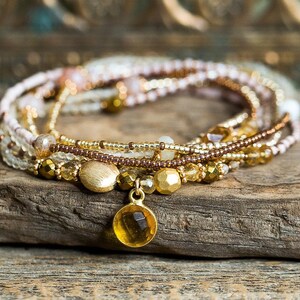 May include: A multi-strand bracelet made of small, gold, brown, and white beads with a gold charm featuring a round, yellow gemstone.