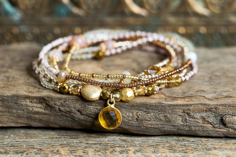 May include: A multi-strand bracelet made of small, gold, brown, and white beads with a gold charm featuring a round, yellow gemstone.