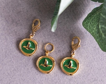 May include: Three gold-colored charms with a green enamel center featuring a gold bird design. Two charms have a lobster clasp, and one has a small ring. The charms are on a gray surface.