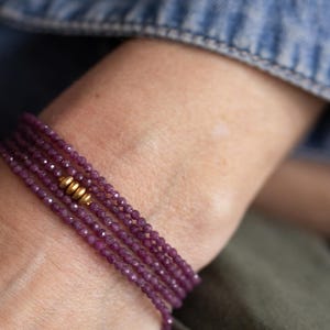 May include: A multi-strand bracelet with small, faceted, deep purple beads. The bracelet is accented with gold-coloured beads. The jewellery is worn on a wrist, with a denim jacket and green fabric in the background.