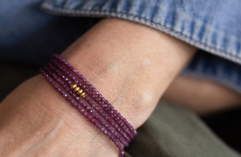 May include: A multi-strand bracelet with small, faceted, deep purple beads. The bracelet is accented with gold-coloured beads. The jewellery is worn on a wrist, with a denim jacket and green fabric in the background.