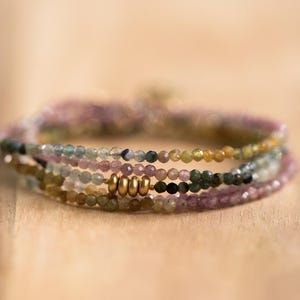 Pu&ograve; includere: Un gruppo di braccialetti di perline nei toni del rosa, verde, giallo e nero. I braccialetti sono realizzati con piccole perline rotonde e presentano dettagli metallici color oro. Sono esposti su una superficie di legno.