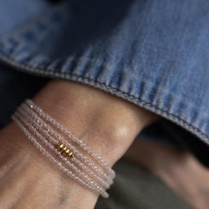 K&ouml;nnte beinhalten: Ein zartes Armband mit mehreren Str&auml;ngen aus kleinen, klaren Perlen. Ein goldfarbenes zylindrisches Akzentst&uuml;ck ist auf dem Armband zentriert. Das Armband wird an einem Handgelenk getragen, mit einer Jeansjacke im Hintergrund.