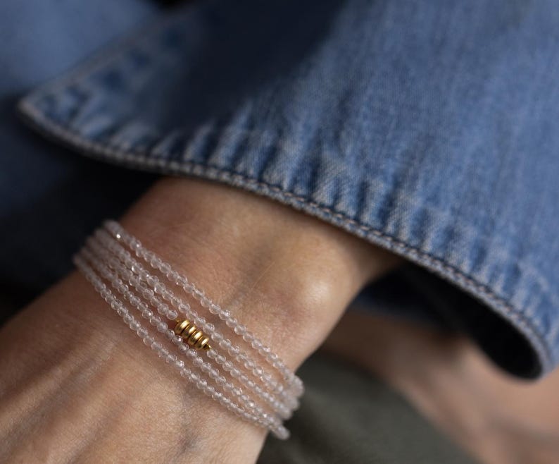 K&ouml;nnte beinhalten: Ein zartes Armband mit mehreren Str&auml;ngen aus kleinen, klaren Perlen. Ein goldfarbenes zylindrisches Akzentst&uuml;ck ist auf dem Armband zentriert. Das Armband wird an einem Handgelenk getragen, mit einer Jeansjacke im Hintergrund.