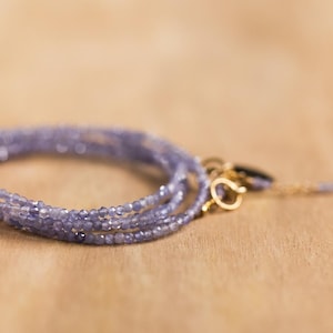 May include: A delicate, multi-strand bracelet featuring small, light purple beads. The bracelet has a gold clasp and a matching gold chain with a small bead at the end. The jewelry is displayed on a light brown wooden surface.