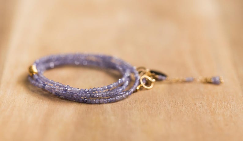 May include: A delicate, multi-strand bracelet featuring small, light purple beads. The bracelet has a gold clasp and a matching gold chain with a small bead at the end. The jewelry is displayed on a light brown wooden surface.