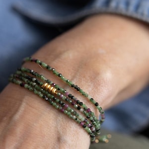 May include: A multi-strand beaded bracelet featuring small, faceted beads in shades of green, black, and pink. The bracelet has a gold-colored accent bead. The jewelry is wrapped around a wrist.