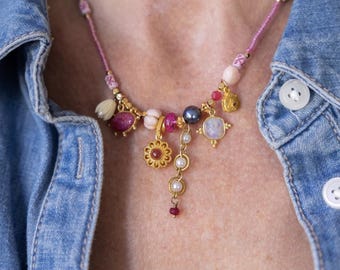 Könnte beinhalten: Eine farbenfrohe Halskette mit verschiedenen Perlen und Anhängern. Die Halskette hat rosa, goldene und perlmuttfarbene Akzente, mit einem zentralen Anhänger in Form einer goldenen Blume. Sie wird über einem Jeanshemd getragen.