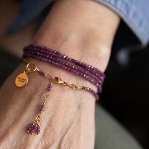 May include: A multi-strand wrap bracelet with small, faceted, deep purple beads. The bracelet has a gold-coloured chain and clasp, with a small gold charm. The charm has text on it. The bracelet is wrapped around a wrist.