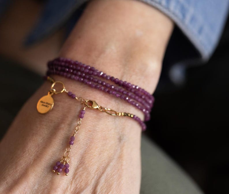 May include: A multi-strand wrap bracelet with small, faceted, deep purple beads. The bracelet has a gold-coloured chain and clasp, with a small gold charm. The charm has text on it. The bracelet is wrapped around a wrist.