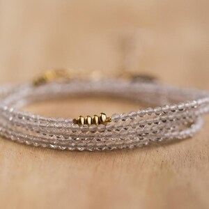K&ouml;nnte beinhalten: Ein zartes Wickelarmband mit mehreren Str&auml;ngen aus klaren, facettierten Perlen. Das Armband ist mit kleinen goldfarbenen Perlen akzentuiert. Der Schmuck wird auf einer hellbraunen Holzoberfl&auml;che pr&auml;sentiert.