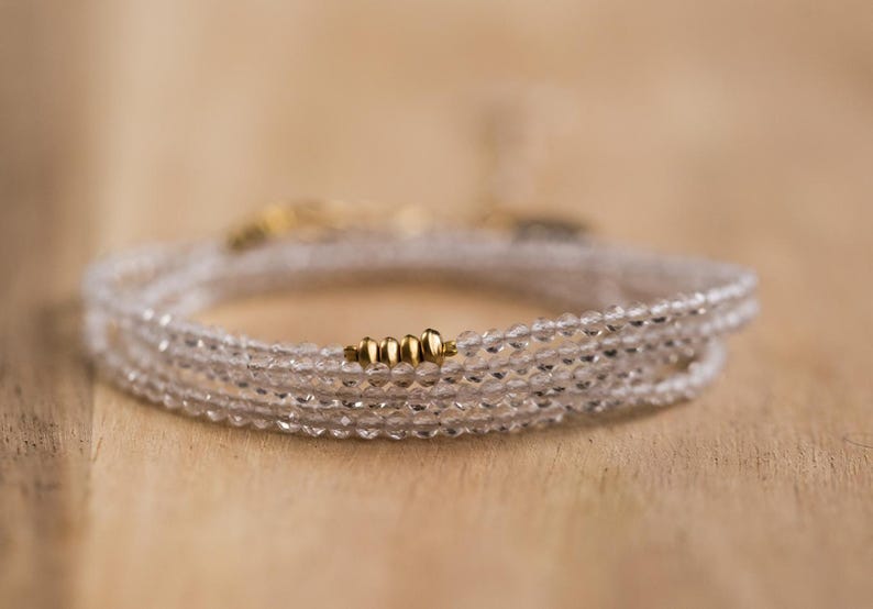 K&ouml;nnte beinhalten: Ein zartes Wickelarmband mit mehreren Str&auml;ngen aus klaren, facettierten Perlen. Das Armband ist mit kleinen goldfarbenen Perlen akzentuiert. Der Schmuck wird auf einer hellbraunen Holzoberfl&auml;che pr&auml;sentiert.