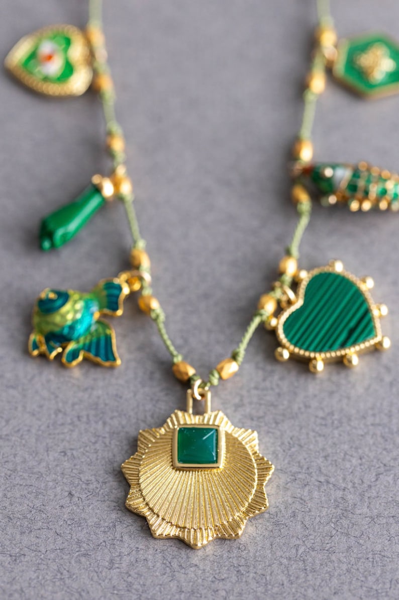 May include: A gold necklace with green gemstone charms. The charms include a heart, a fish, a star, and a small green stick. The necklace is made of a thin, green cord.