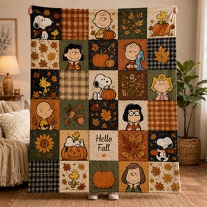 May include: A rectangular throw blanket featuring a patchwork design of Peanuts characters, pumpkins, sunflowers, and autumn leaves in orange, brown, green, and black. The center square displays the text Hello Fall with a small heart underneath.