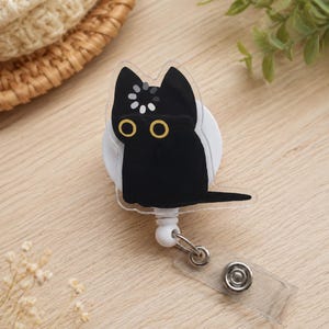 May include: A badge reel featuring a clear acrylic cutout of a black cat with large yellow eyes and a white loading icon on its forehead. The cat is attached to a white circular base with a retractable cord, a metal ring, and a clear plastic strap.
