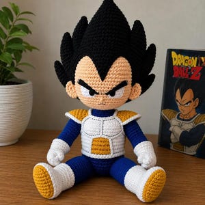 Op de afbeelding: Een gehaakte pop van het personage Vegeta uit Dragon Ball Z, met een groot zwart kapsel, een getinte huid en een blauw-wit pak met gele harnasaccenten. De pop heeft een strenge gezichtsuitdrukking en zit rechtop.