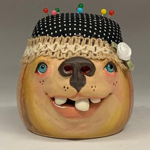 Puede incluir: Un alfiletero caprichoso con forma de cabeza de perro con un sombrero de lunares negros y blancos, una cinta de crochet marrón y una flor blanca. El perro tiene ojos azules, una nariz marrón y una sonrisa  dentada.
