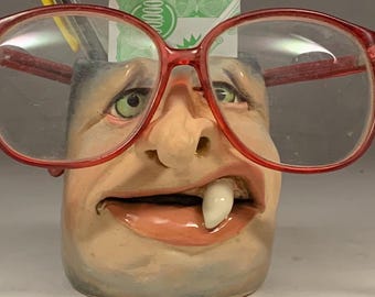 May include: A whimsical ceramic pencil holder shaped like a face, complete with a large tooth, red-framed glasses, and pens. The holder has a light skin tone and is filled with pens and a small piece of paper.