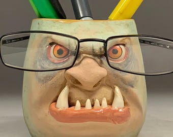 May include: A unique, hand-painted pen holder shaped like a monster's face, with orange eyes, large teeth, and black-framed glasses. The holder contains a green, black, and yellow marker and a gray pen. The text "The Art of Lucky Stradley" is at the bottom.