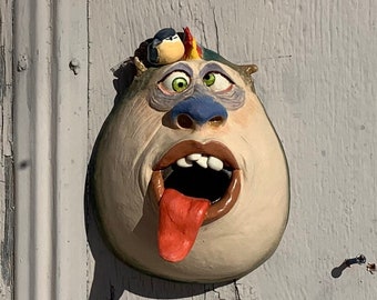 Può includere: Una scultura in ceramica fantasiosa di un viso con la bocca spalancata e una lunga lingua che sporge. La scultura è dipinta in tonalità di blu, verde e rosso, e ha un piccolo uccello appollaiato sulla testa.