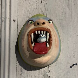 以下が含まれることがあります： 口を開けて小さな灰色の生き物を中に見せている、モンスターの頭部のセラミック彫刻。モンスターは緑、青、白で塗られた顔と赤い舌をしています。
