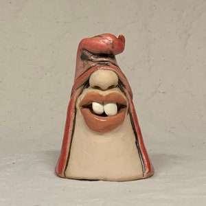 May include: A small ceramic sculpture of a face with a wide smile and large teeth. The sculpture is painted in shades of pink, brown, and white. The face is wearing a red hat.
