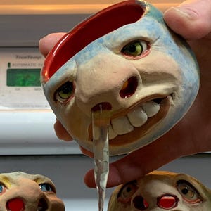 May include: A collection of whimsical, face-shaped ceramic bowls and cups. One cup is pouring a liquid into a larger bowl. The bowls and cups have expressive features, including eyes, noses, and mouths. An eggshell is also present.