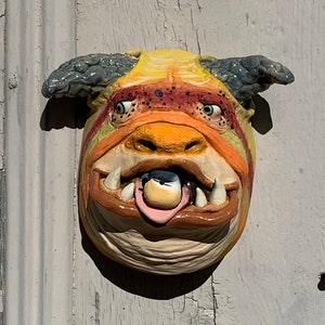 Könnte beinhalten: Ein gelber, orangefarbener und schwarzer Keramikmonsterkopf mit einem großen, offenen Maul und einer herausgestreckten Zunge. Das Monster hat ein kleines, rundes, schwarzes Auge.