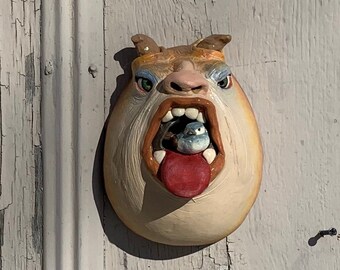Può includere: Una scultura in ceramica di un bulldog con una grande bocca aperta, dipinta in tonalità di marrone, bianco e blu. Il bulldog tiene un piccolo oggetto blu in bocca.