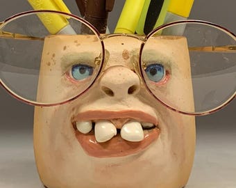 May include: A whimsical pen holder shaped like a face, with blue eyes, a wide grin, and round eyeglasses. The holder contains several pens and markers in shades of yellow and brown. The text "The Art of Lucky Stradley" is at the bottom.