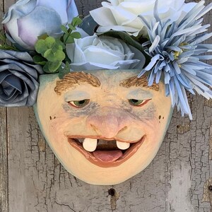 May include: A whimsical ceramic planter shaped like a face with a toothy grin, decorated with blue and white flowers. The planter is a unique and quirky addition to any home decor.