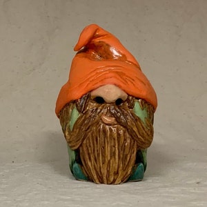 Può includere: Una piccola statuetta di gnomo in ceramica con un cappello arancione, una barba marrone e vestiti verdi. Lo gnomo è seduto con le mani giunte davanti al viso.