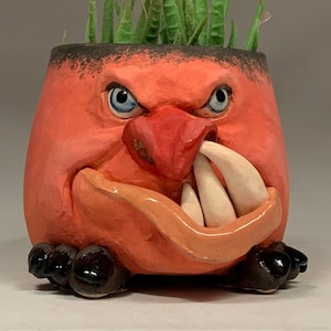 May include: A red ceramic planter shaped like a face with a toothy grin and a green plant growing out of the top. The planter has black eyes and a red nose.