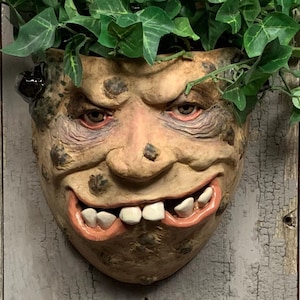 May include: A grotesque, face-shaped planter with a wide, toothy grin and green foliage sprouting from the top. The planter is painted in shades of tan and brown, with a textured surface. The text "The Art of Lucky Stradley" is at the bottom.