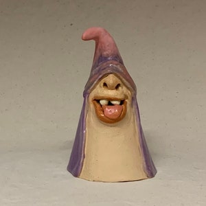 Può includere: Una statuetta in ceramica fantasiosa di un personaggio con un cappello viola e rosa, un ampio sorriso e una lingua che sporge. La statuetta è realizzata in argilla e ha una superficie testurizzata.