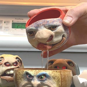 May include: A collection of whimsical, face-shaped ceramic bowls and sculptures. One bowl is held, pouring a liquid into another bowl. The sculptures have exaggerated features and expressions, with a cracked eggshell nearby. The text "The Art of Lucky Stradley" is at the bottom.