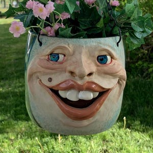 Puede incluir: Una maceta colgante caprichosa con un diseño de cara sonriente, con ojos azules, labios grandes y un lunar. La maceta está llena de flores rosas y follaje verde, suspendida por una cadena negra. El texto "The Art of Lucky Stradley" está en la parte inferior.