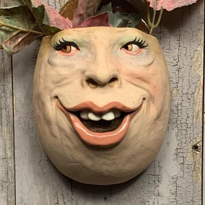 May include: A whimsical, wall-mounted planter shaped like a smiling face. The beige ceramic face has detailed features, including painted eyelashes, rosy cheeks, and open lips revealing teeth. The planter holds faux greenery with pink and green leaves. The text "The Art of Lucky Stradley" is at the bottom.