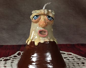 May include: A ceramic candle holder shaped like a person's head with a dripping wax effect. The candle holder is brown with a yellow face and blue eyes. The candle holder is sitting on a white crocheted doily.
