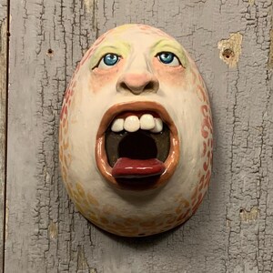 Puede incluir: Una escultura de cerámica de una cara con la boca abierta y una lengua roja. La cara tiene forma de huevo con ojos azules, dientes blancos y una superficie texturizada y moteada. El texto "The Art of Lucky Stradley" está en la parte inferior.