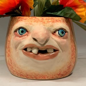 以下が含まれることがあります： 青い目、鼻、歯のある口が特徴の、顔のデザインが施された気まぐれなセラミックプランター。プランターには、鮮やかなオレンジと黄色の花と、緑の葉と長い緑の草が入っています。「The Art of Lucky Stradley」というテキストが一番下にあります。