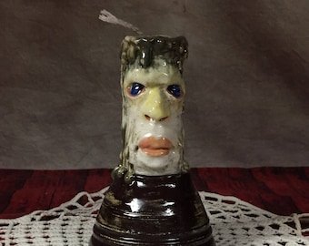May include: A ceramic candle holder shaped like a face with dripping wax. The face has a light brown skin tone, blue eyes, a yellow nose, and a pink mouth. The candle holder is sitting on a white crocheted doily.
