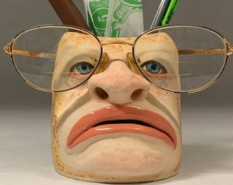 May include: A ceramic pencil holder shaped like a face with a sad expression. The face has a large nose, brown eyebrows, and blue eyes. The holder is wearing gold-rimmed glasses and has a brown, green, and white pen and pencil inside.